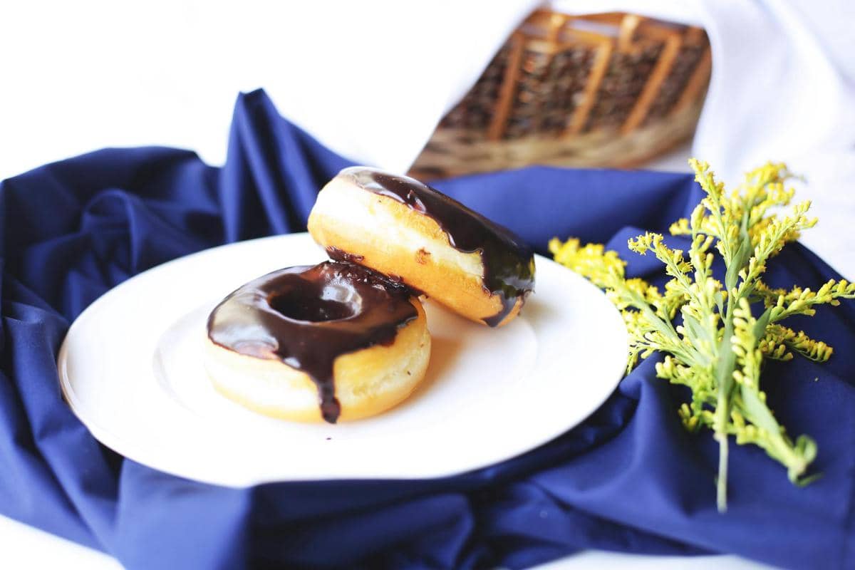 Dua donat dengan lapisan cokelat mengilap di atas piring putih, diletakkan di atas kain biru dengan hiasan bunga kuning di sampingnya.