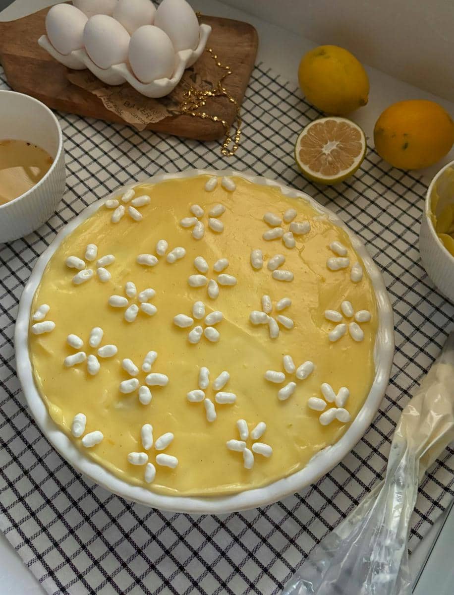 Lemon tiramisu berlapis krim kuning dihiasi motif bunga putih, dikelilingi bahan seperti telur, lemon, dan kopi di atas kain kotak.
