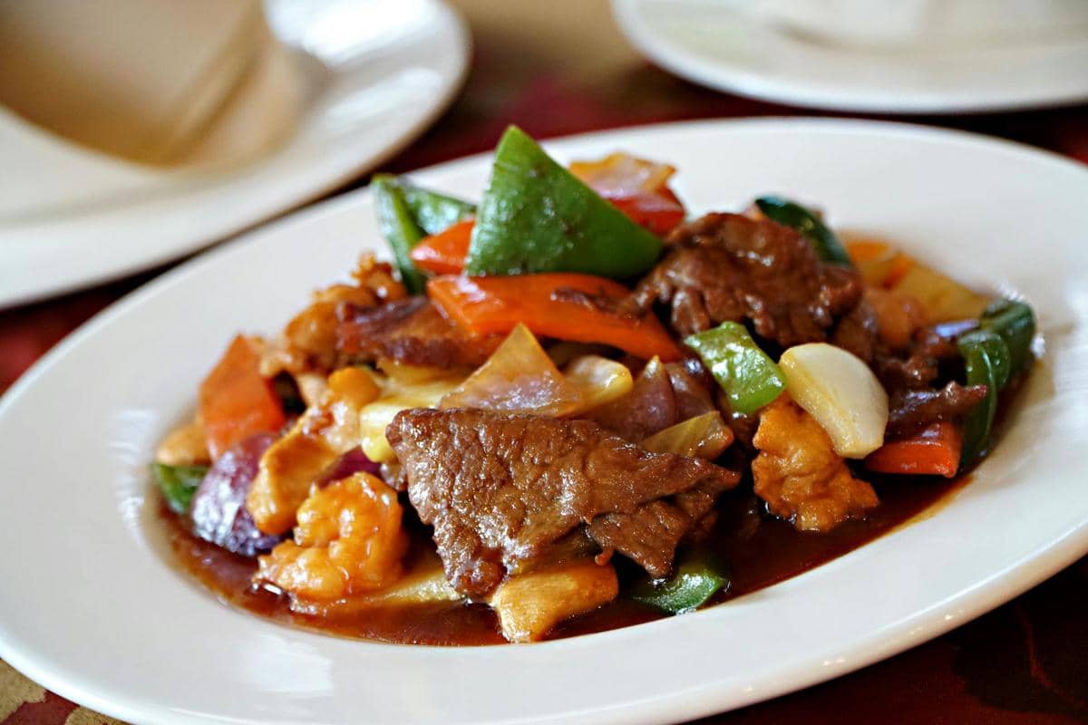Hidangan olahan daging kambing dengan saus cokelat, disajikan bersama potongan paprika, bawang, dan sayuran di atas piring putih.