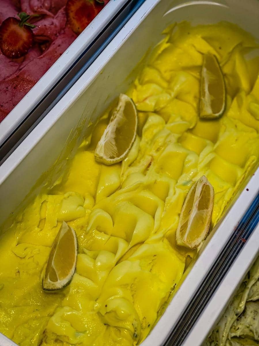 Es krim lemon berwarna kuning cerah dalam wadah logam dengan potongan lemon di atasnya, berdampingan dengan es krim rasa lain.