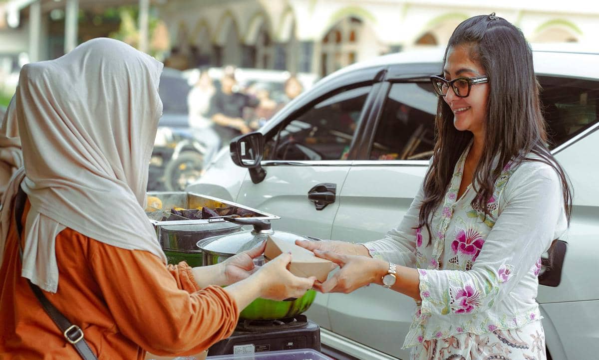 Dua perempuan saling memberikan makanan di luar ruangan, salah satunya mengenakan hijab dan satunya tersenyum sambil mengenakan kacamata.