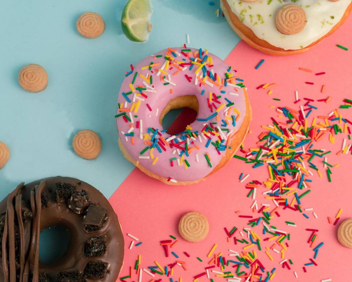 Beberapa donat berwarna-warni dengan topping cokelat, krim, dan taburan sprinkle di atas permukaan dua warna biru dan merah muda.