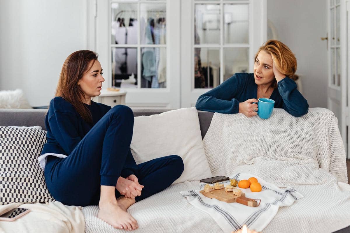 Dua perempuan duduk di sofa ruang tamu sedang mengobrol santai, satu memegang cangkir biru dan yang lain duduk bersila mengenakan pakaian santai.