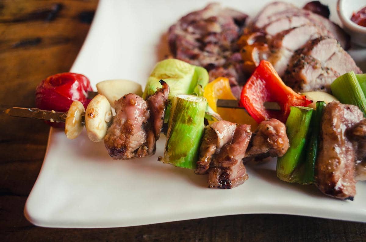 Sate daging dengan potongan paprika, bawang, dan sayuran berwarna cerah tersaji di piring putih di atas meja kayu.