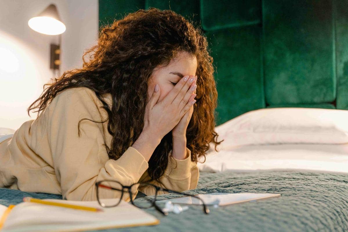 Seorang perempuan berambut keriting menutupi wajah dengan tangan sambil berbaring di tempat tidur, di depannya terdapat buku dan kacamata.