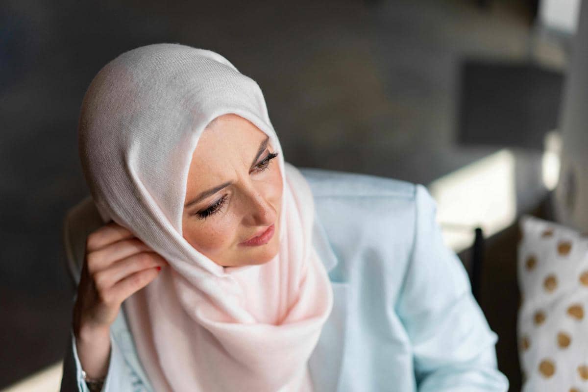 Perempuan berhijab berwarna putih dan pastel duduk sambil menyentuh hijabnya dengan ekspresi tenang di dalam ruangan.