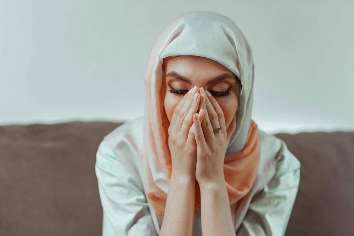 Seorang perempuan berhijab duduk dengan tangan menutupi wajah, tampak sedang merenung atau merasa sedih di atas sofa.