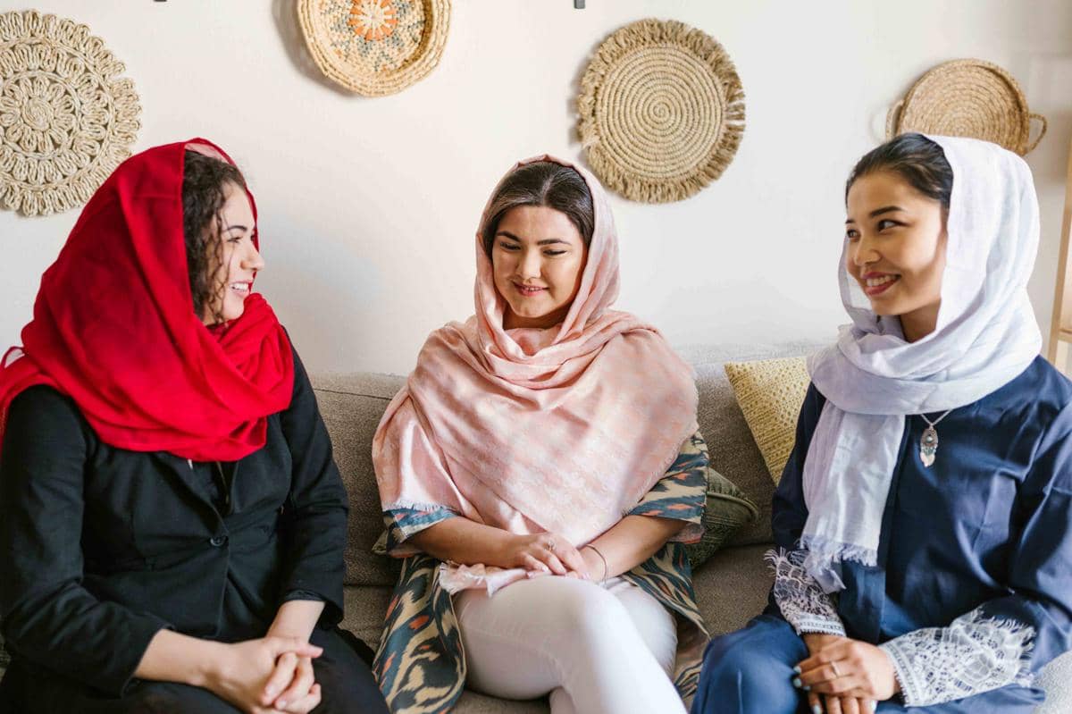 Tiga wanita berhijab duduk di sofa sambil tersenyum dan berbincang di ruang tamu dengan hiasan dinding anyaman rotan.