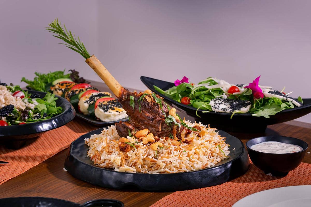Nasi kebuli dengan daging kambing bertulang disajikan di atas piring hitam, dihiasi kacang dan bawang goreng, dengan lauk dan salad di sekitarnya.