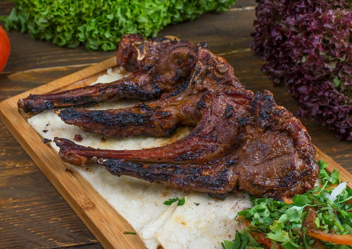 Potongan daging kambing panggang disajikan di atas talenan kayu dengan roti pipih, sayuran segar, dan daun selada di sekitarnya.