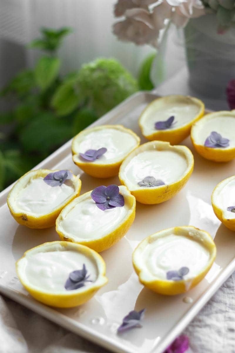 Beberapa lemon posset disajikan dalam kulit lemon setengah, dihiasi bunga ungu kecil di atas nampan putih dengan latar tanaman hijau.