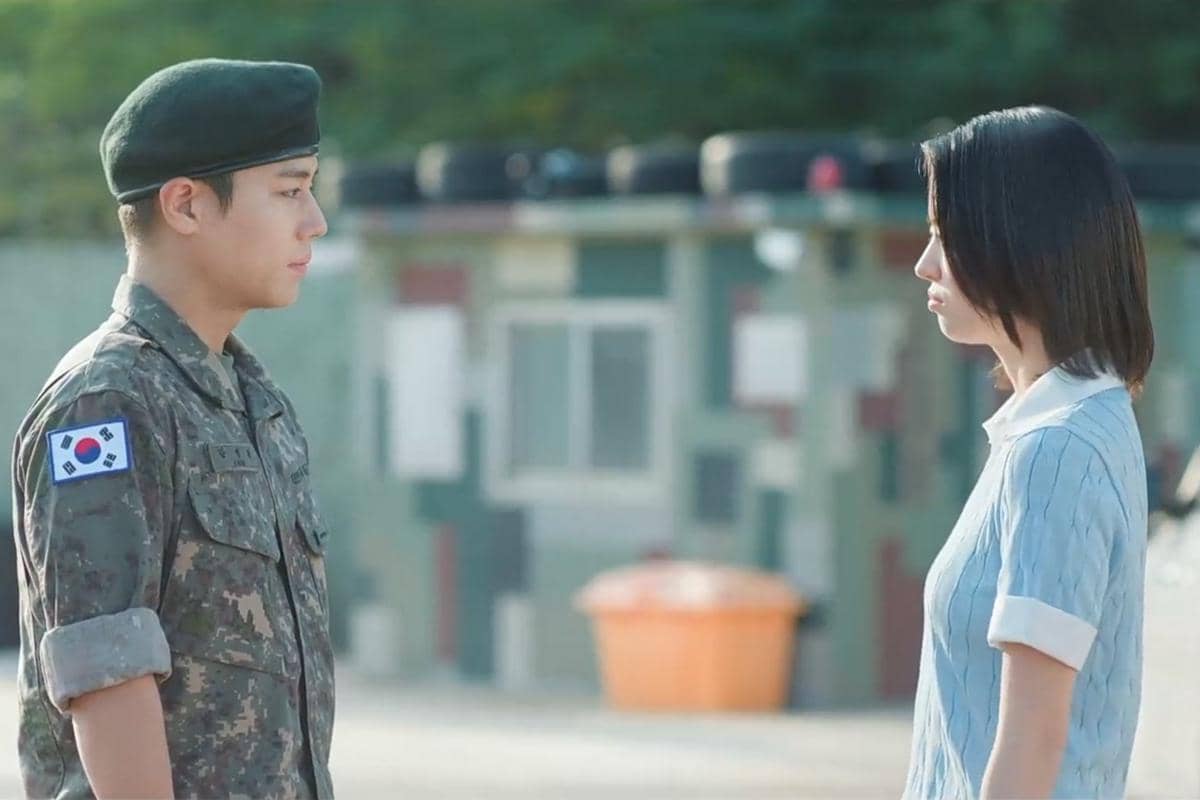 Seorang tentara berseragam militer Korea berdiri berhadapan dengan seorang wanita berambut pendek mengenakan pakaian biru muda di luar ruangan.