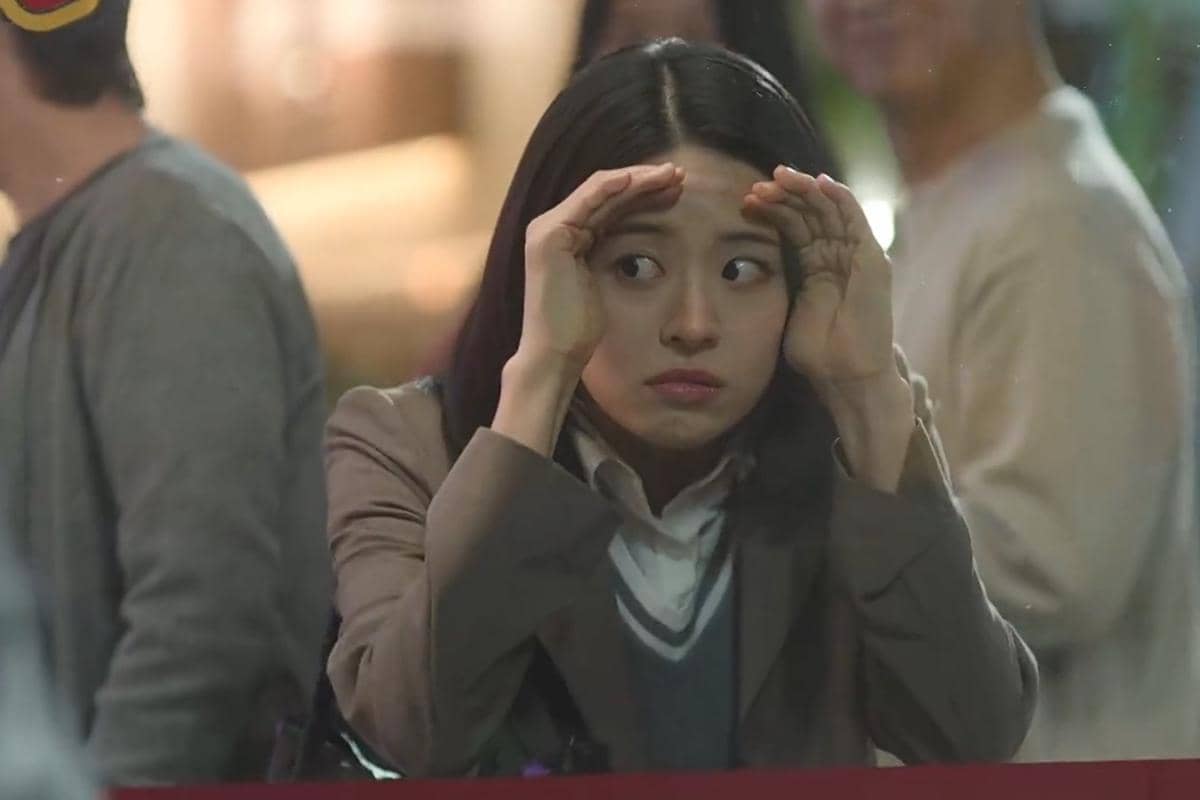 Seorang perempuan mengenakan jas cokelat dan kemeja putih tampak mengintip melalui jendela dengan ekspresi penasaran di adegan drama The Legend of Kitchen Soldier.