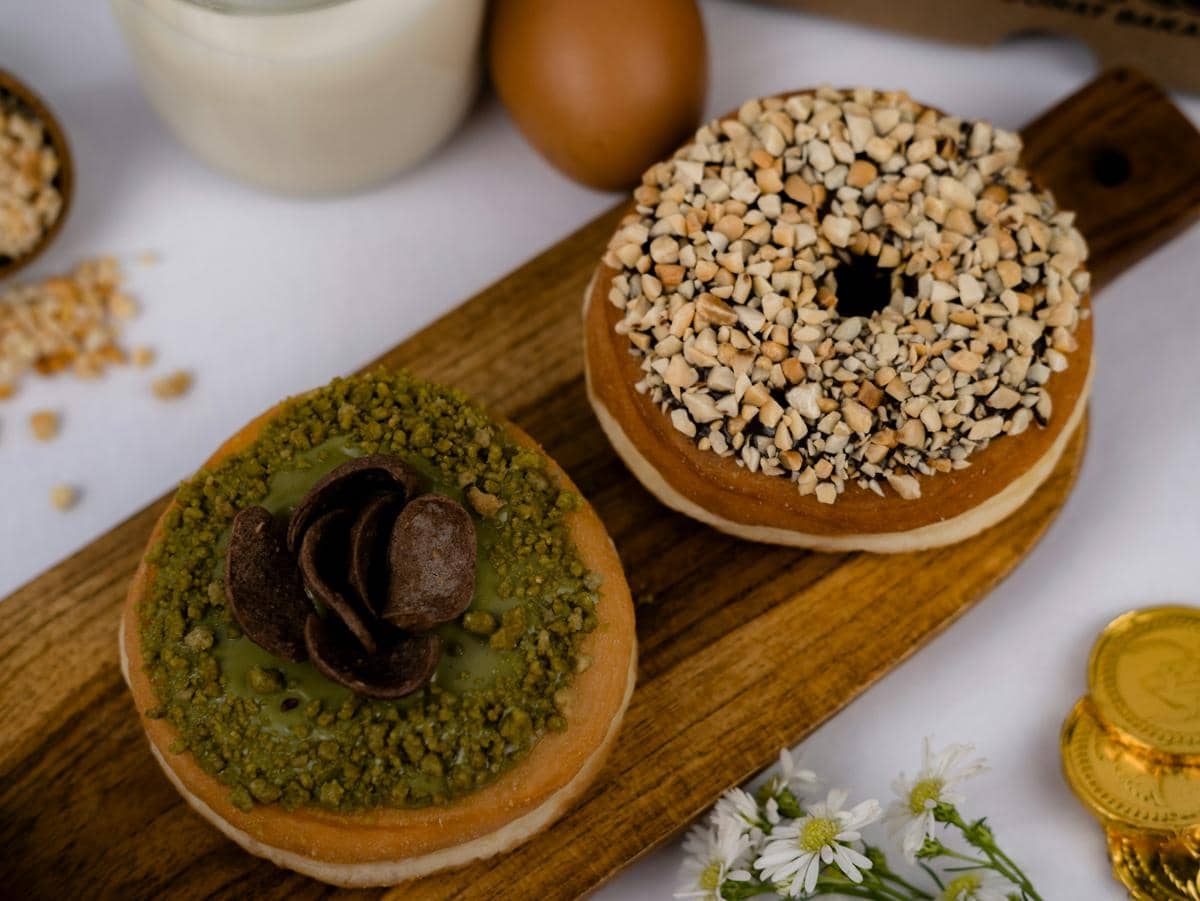 Dua donat dengan topping kacang cincang dan krim hijau matcha di atas papan kayu, disajikan bersama susu dan telur di latar belakang.