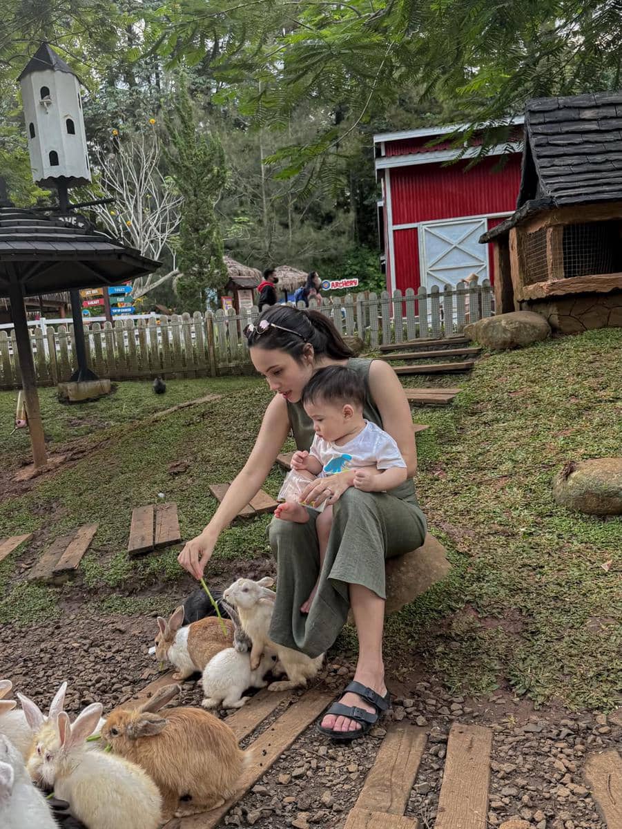 Seorang wanita duduk sambil memangku anak kecil dan memberi makan beberapa kelinci di area taman dengan suasana alami.