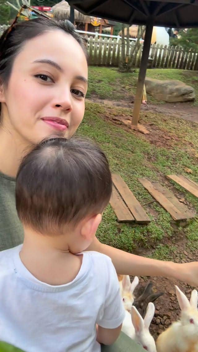 Seorang wanita menggendong anak kecil sambil memberi makan beberapa kelinci di area taman dengan rumput dan pagar kayu.