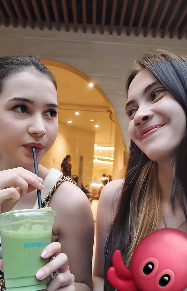 Dua remaja perempuan tersenyum sambil berbincang santai, salah satunya memegang minuman hijau di gelas plastik dengan sedotan hitam.