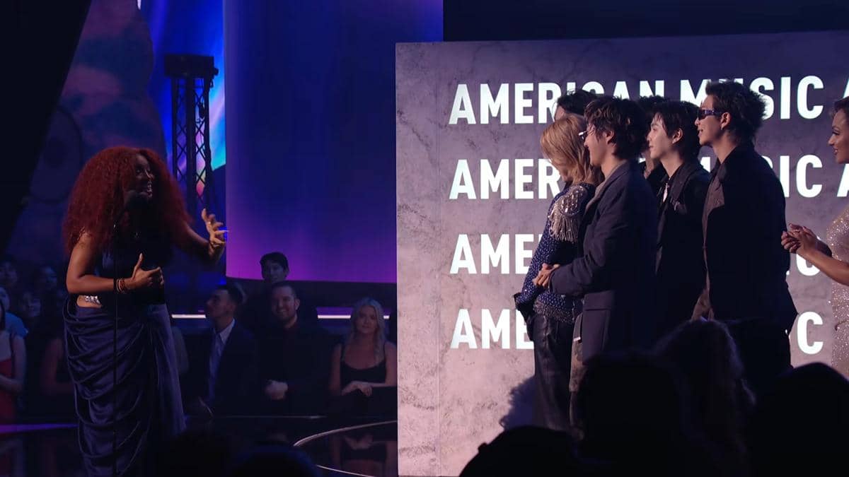 Seorang penyanyi bergaun biru berbicara di panggung kepada sekelompok pria berpakaian formal di acara American Music Awards 2026.