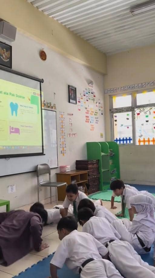 Seorang guru bersama murid kelas 1 SD melakukan latihan push-up di ruang kelas dengan presentasi edukasi kesehatan di layar proyektor.