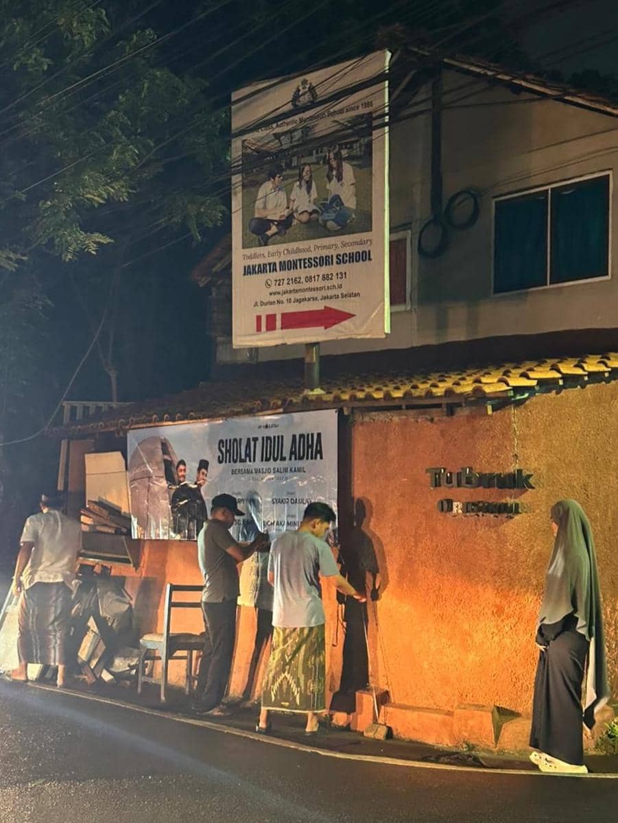 Beberapa orang menyiapkan area di depan tembok dengan spanduk bertuliskan Sholat Idul Adha pada malam hari di Jakarta Selatan.
