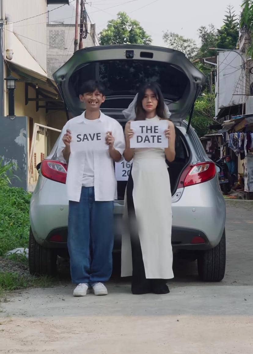 Sepasang pria dan wanita berdiri di depan mobil dengan bagasi terbuka sambil memegang kertas bertuliskan 'Save the Date'.