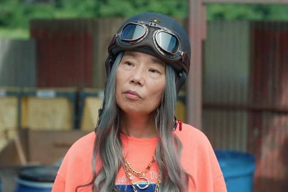 Seorang wanita berambut panjang abu-abu mengenakan helm motor dan kacamata pelindung di atas kepala, berpakaian kasual dengan kaus oranye.