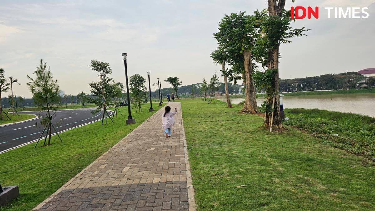 Seorang anak berlari di jalur pedestrian tepi Danau Cihuni, Tangerang, dengan pemandangan hijau dan jalan berlampu di sisi kiri.