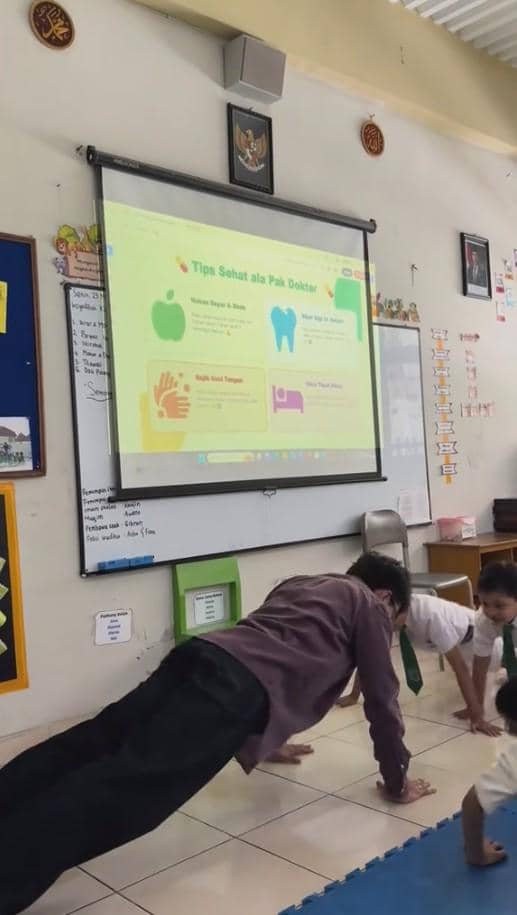 Seorang pria dewasa dan beberapa siswa SD melakukan push-up di ruang kelas dengan layar proyektor berisi tips hidup sehat.