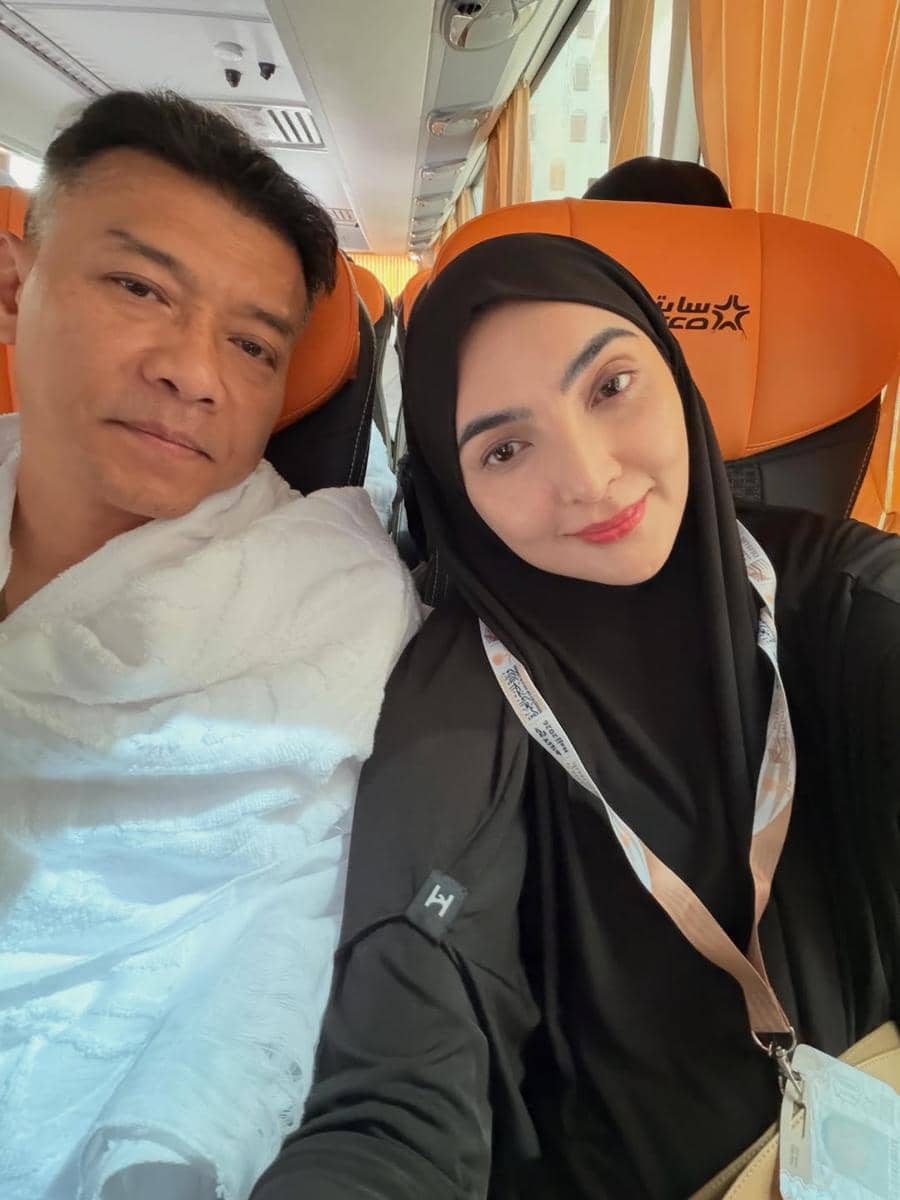 Seorang pria dan wanita berpose bersama di dalam bus dengan kursi berwarna oranye, wanita mengenakan hijab hitam dan pria memakai kain putih.