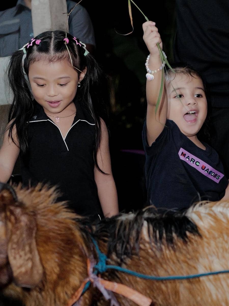 Dua anak kecil tersenyum sambil melihat seekor kambing kurban, salah satunya mengangkat rumput di tangannya dengan gembira.