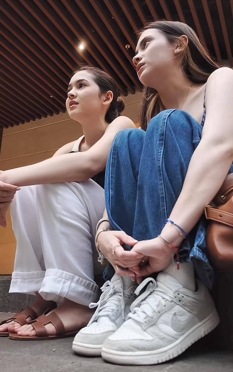 Dua perempuan duduk santai di lantai dengan gaya kasual, satu mengenakan celana putih dan sandal, satunya memakai jeans dan sepatu putih.