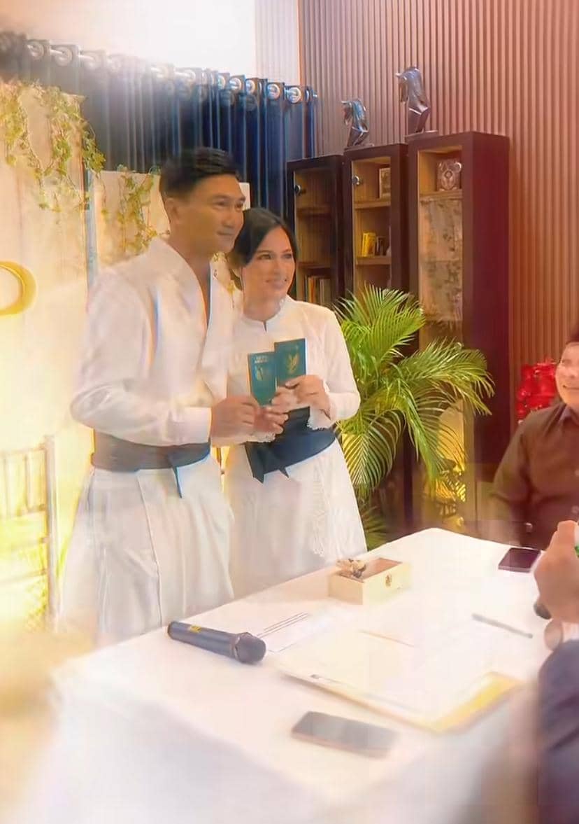 Sepasang pengantin mengenakan busana putih memegang buku nikah di sebuah ruangan dengan dekorasi sederhana dan tanaman hias.