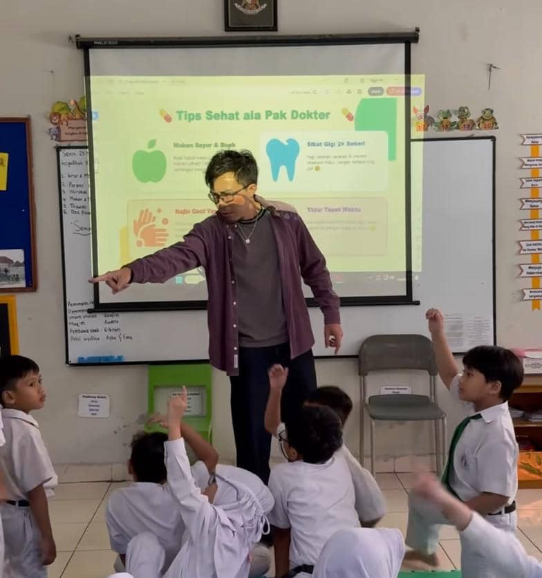 Seorang pria mengajar siswa kelas 1 SD di depan layar proyektor yang menampilkan materi tentang tips hidup sehat di ruang kelas.