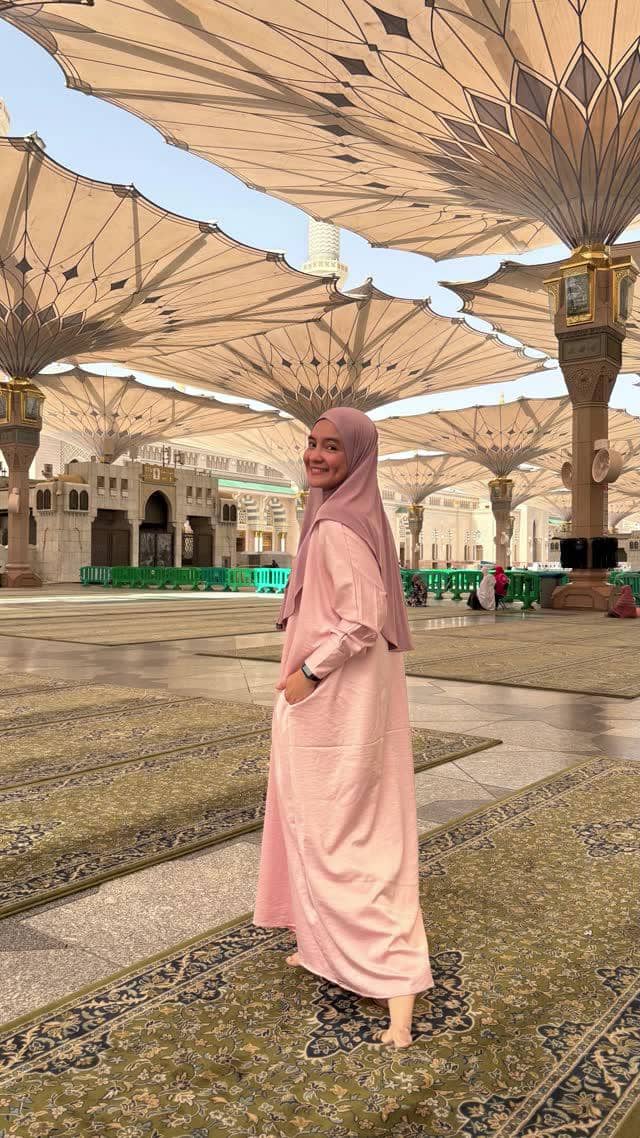 Seorang wanita berhijab berwarna ungu muda mengenakan gamis pink berdiri tersenyum di area Masjid Nabawi dengan payung besar terbuka.