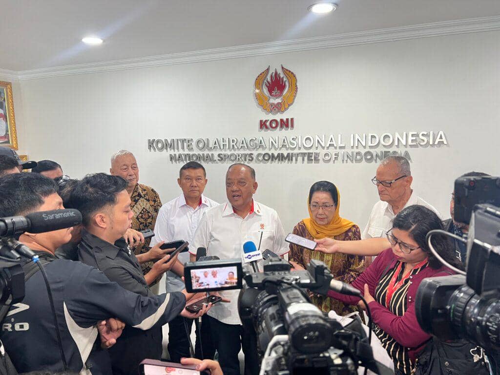 Beberapa perwakilan KONI memberikan keterangan pers di depan logo Komite Olahraga Nasional Indonesia dengan sejumlah wartawan dan kamera merekam.