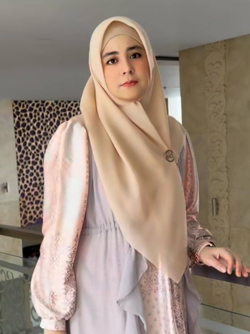Seorang wanita berhijab krem mengenakan gaun panjang berwarna pastel berdiri di dalam ruangan dengan latar dinding bermotif.