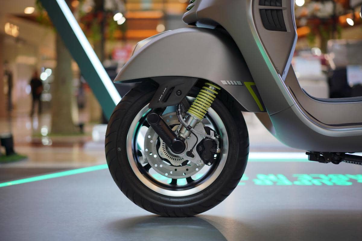 Roda depan Vespa GTS Super Tech 250 berwarna abu-abu dengan suspensi kuning dan cakram rem terlihat jelas di area pameran dalam ruangan.
