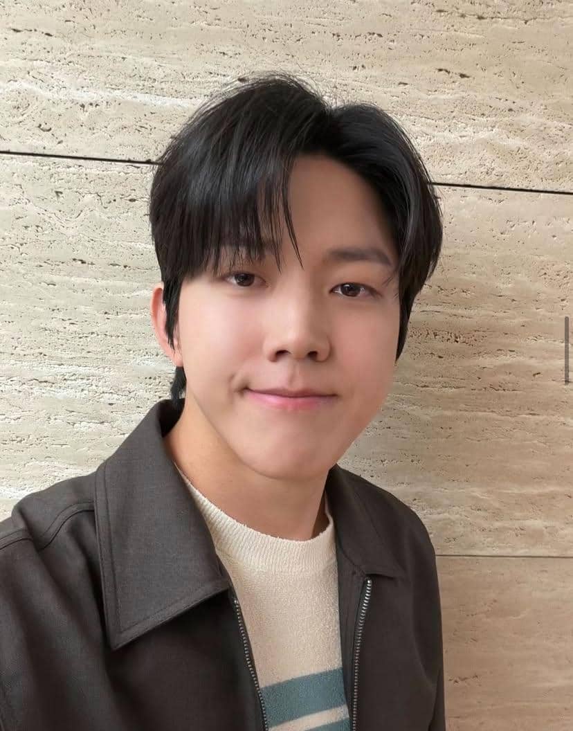 Seorang pria berambut hitam tersenyum tipis mengenakan jaket cokelat dan sweater krem bergaris biru di depan dinding batu krem.