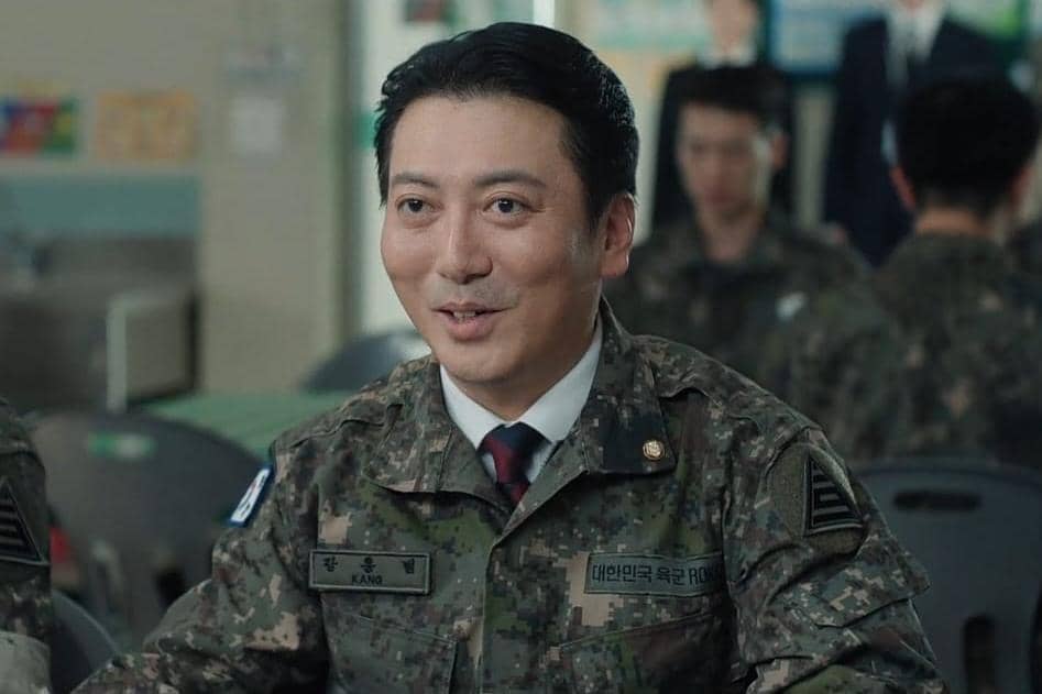 Seorang tentara berpakaian seragam militer sedang duduk dan tersenyum di ruang makan dalam adegan drama Korea The Legend of Kitchen Soldier.