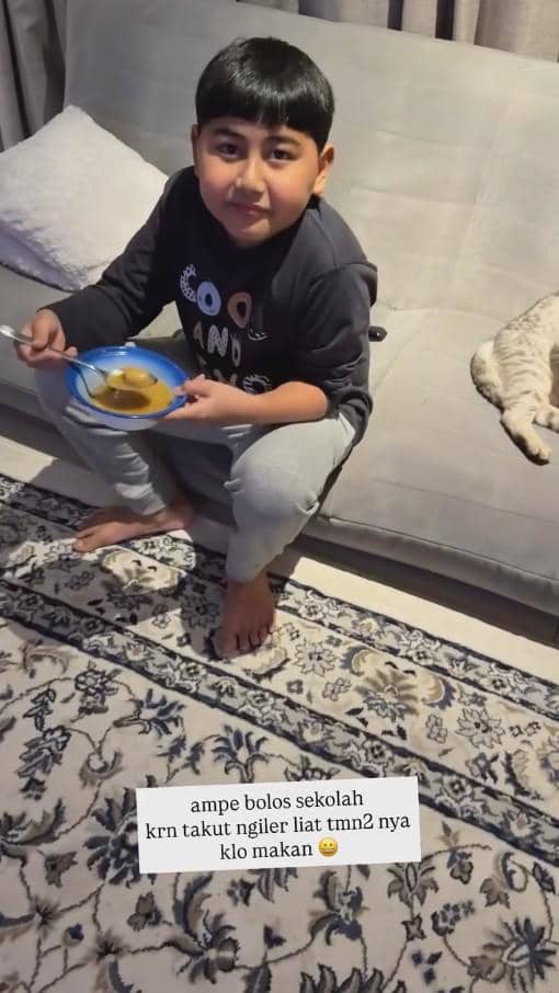 Seorang anak laki-laki duduk di sofa sambil memegang mangkuk berisi makanan untuk berbuka puasa, dengan seekor kucing berbaring di sampingnya.