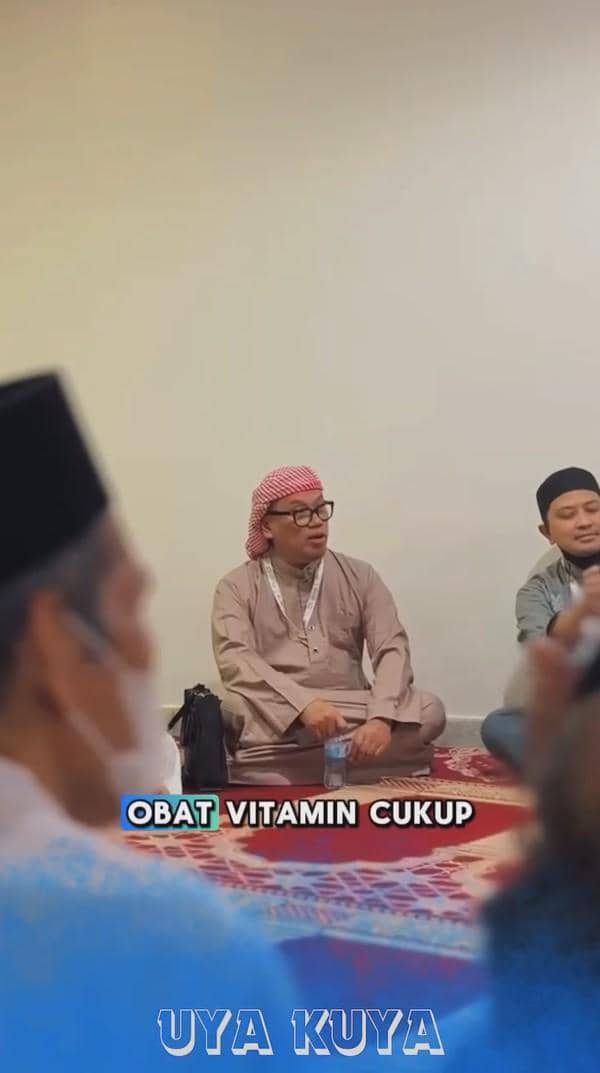 Seorang pria mengenakan gamis cokelat dan sorban merah putih duduk bersila di atas karpet bersama beberapa orang di ruangan.