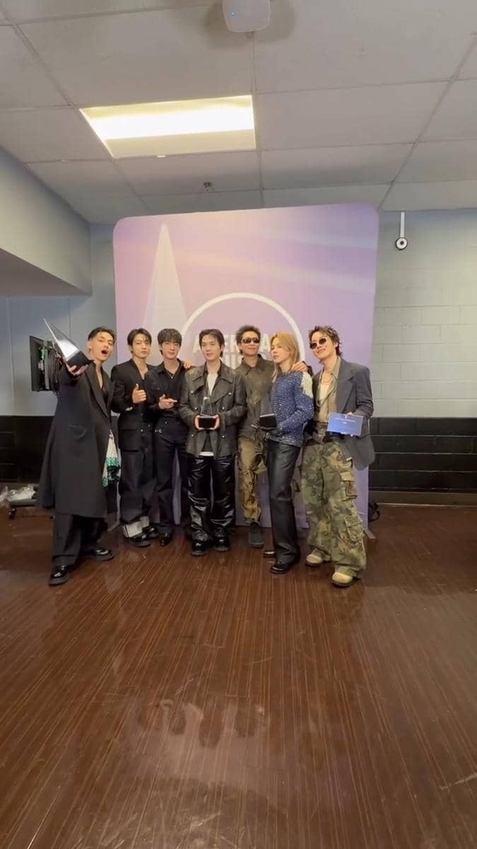Tujuh anggota grup musik berdiri bersama di depan latar bertema American Music Awards 2026 sambil memegang piala penghargaan.