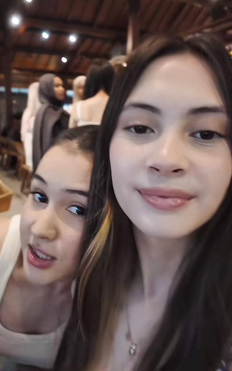 Dua remaja perempuan berpose bersama di tempat makan dengan latar belakang beberapa orang yang sedang berbincang.
