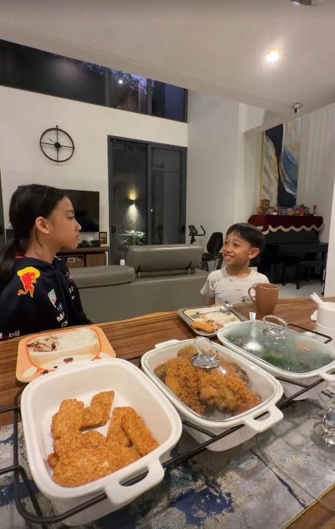 Dua anak duduk di meja makan dengan hidangan tempe goreng, ayam goreng, dan sayur saat berbuka puasa di ruang makan rumah.