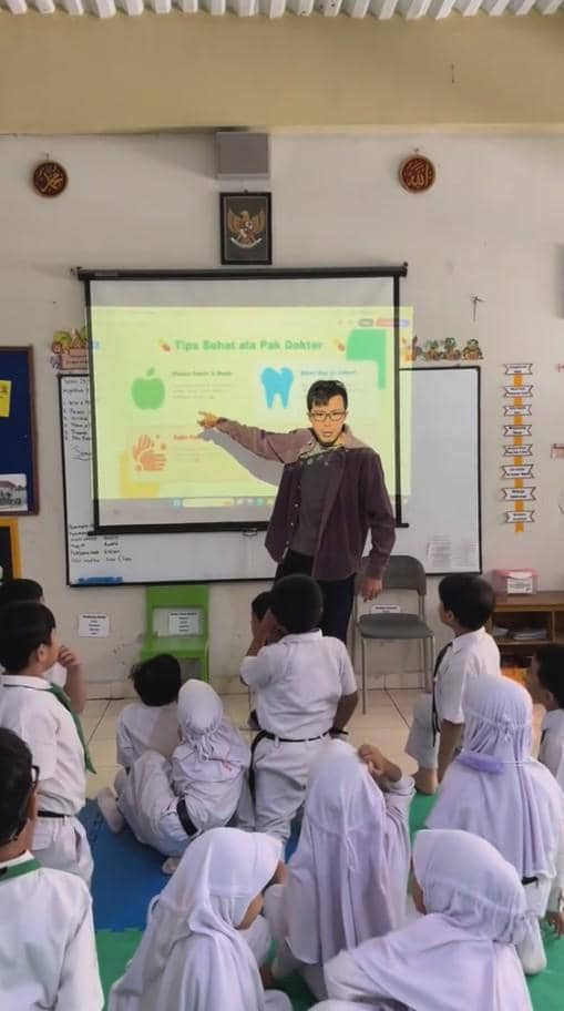 Seorang pria berdiri di depan kelas SD sambil menunjuk presentasi tentang tips sehat, di hadapan murid-murid yang duduk memperhatikan.