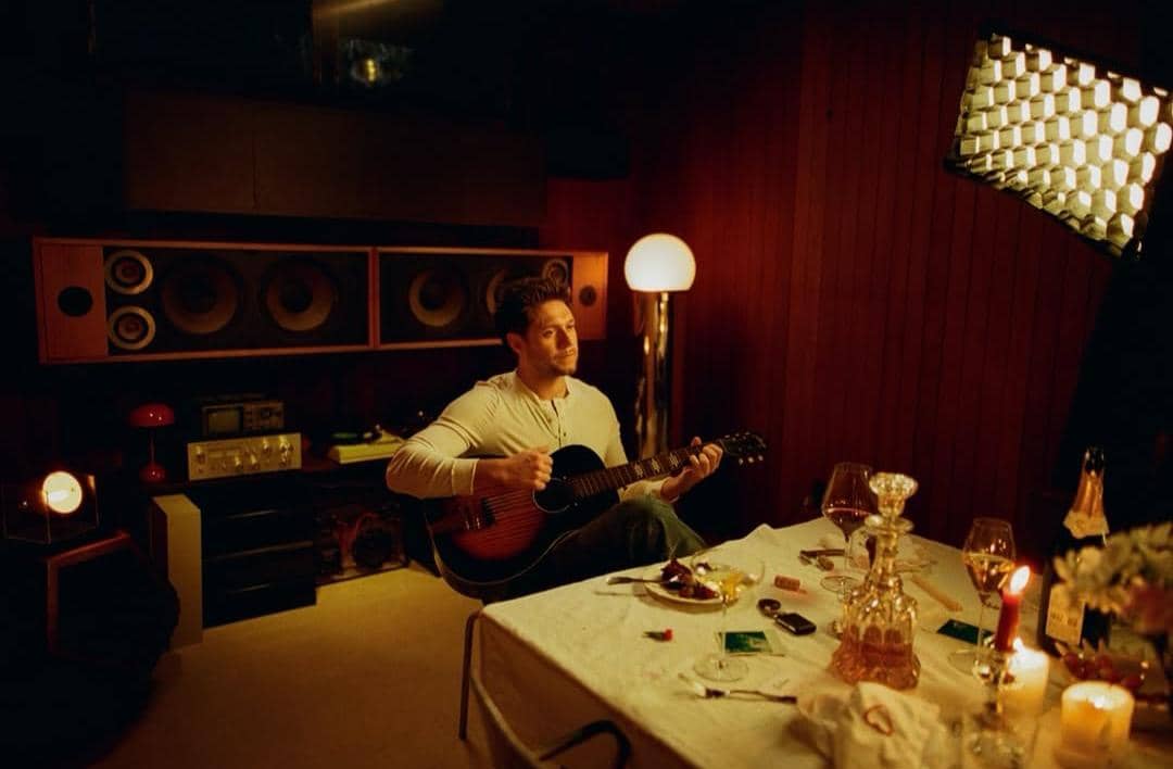 Seorang pria duduk di ruangan bernuansa hangat sambil memainkan gitar akustik di dekat meja makan dengan lilin dan gelas anggur.