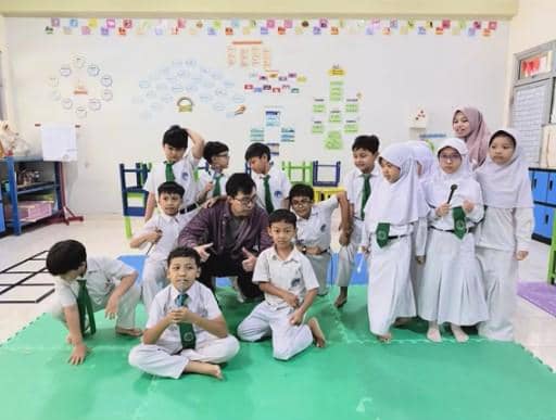 Seorang pria duduk di tengah sekelompok murid sekolah dasar yang mengenakan seragam putih dan dasi hijau di ruang kelas berwarna cerah.