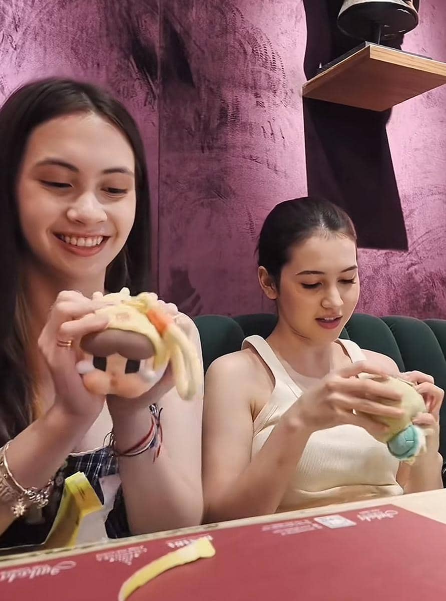 Dua perempuan muda duduk di kafe dengan dinding ungu, tersenyum sambil memegang boneka kecil di tangan mereka.