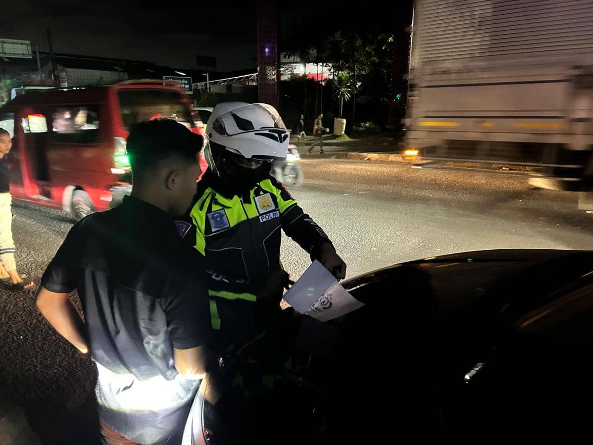 Polisi lalu lintas mengenakan helm dan rompi reflektif sedang menilang pengemudi mobil di jalan kawasan Cikupa pada malam hari.
