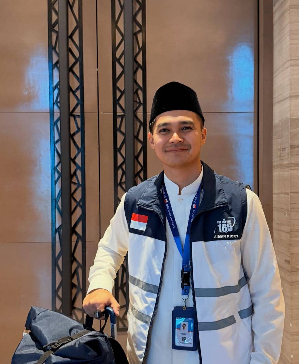 Seorang pria mengenakan peci hitam, rompi bertanda bendera Indonesia, dan membawa tas di dalam ruangan dengan latar dinding modern.